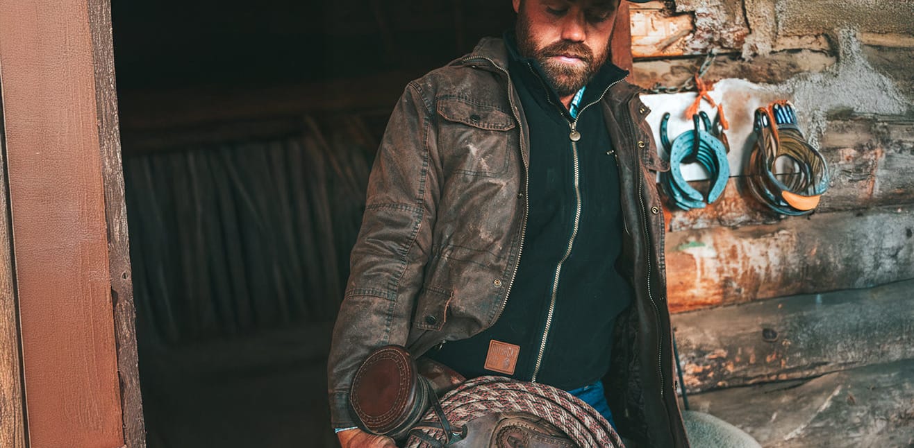 A man wearing a distressed brown leather jacket, black zip-up hoodie, and cowboy hat stands in a rustic log cabin doorway holding a leather saddle.