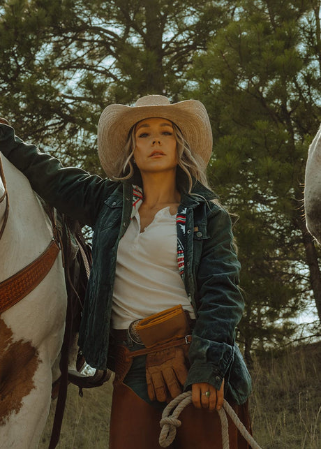 Woman in cowboy hat and leather jacket standing next to a horse with trees in the background